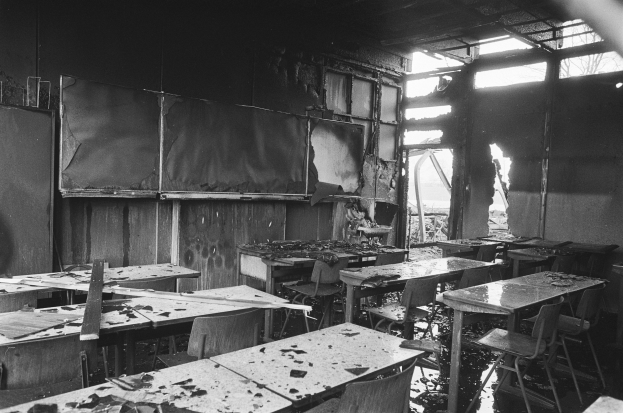 Schwarz-weiß-Foto eines Klassenzimmers mit verkohlten Wänden, zerbrochenen Fenstern und verstreuten Tischen und Stühlen.