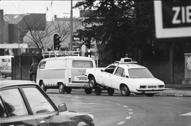 Schwarzes und weißes Foto eines Autos, das von einem Polizeiwagen auf einer Stadtstraße geschleppt wird, mit anderen Fahrzeugen, einem Fußgänger, Ampeln, Pfählen, Bäumen, Gebäuden und einem Zaun im Hintergrund.