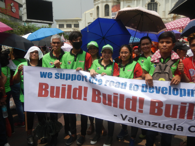 Eine Gruppe von Menschen mit Schirmen und einem Banner mit der Aufschrift "Wir unterstützen den Weg zur Entwicklung von Bau", die auf einer Straße mit Gebäuden, Bäumen und einem klaren blauen Himmel im Hintergrund stehen.