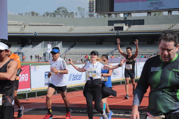 Gruppe von Menschen, die auf einer Stadionbahn laufen, mit Bannern, einem Display, einem Gebäude, Bäumen und einem bewölkten Himmel; einige Läufer tragen Sneakers und Hüte.