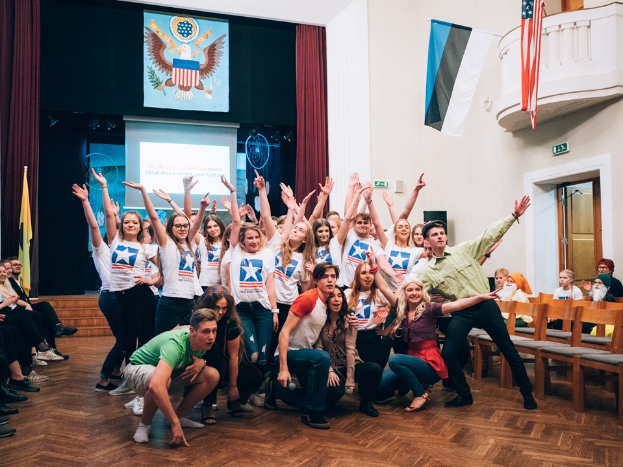 Gruppe von Menschen, die vor einem sitzenden und stehenden Publikum auf einem nationalen Jugendkongress posen, mit einem Bildschirm, Flaggen und anderen veranstaltungsbezogenen Elementen im Hintergrund.