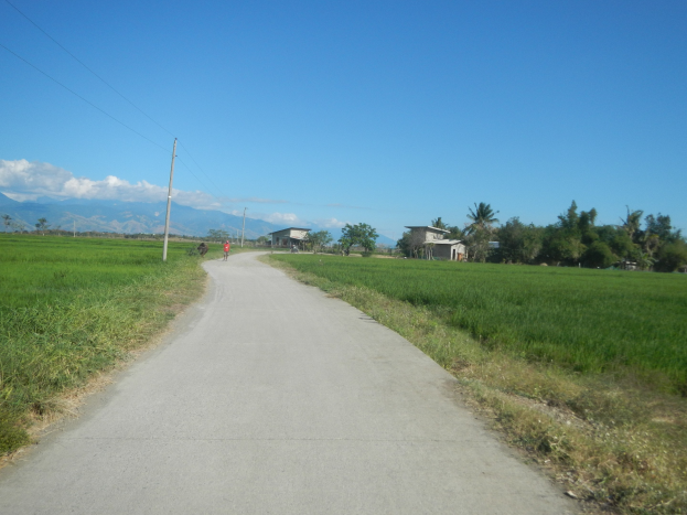 Ein Schotterweg schlängelt sich durch ein grünes Feld mit Fahrradfahrern, Strommasten mit Drähten, Häusern, Bäumen, Hügeln und einem bewölkten Himmel im Hintergrund.