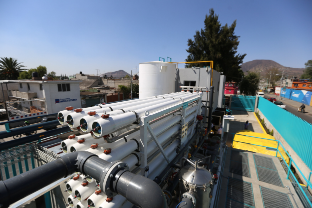 Große Gruppe von Industrieleitungen, Maschinen und Geräten auf einem Gebäudedach in einer städtischen Umgebung mit Straßen, Fahrzeugen, Menschen, Gebäuden, Bäumen, Strommasten, fernen Hügeln und einem klaren blauen Himmel.