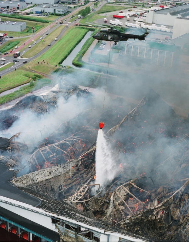 Hubschrauber fliegt über ein großes, durch Feuer zerstörtes Gebäude mit qualmendem Rauch, umgeben von Gebäuden, Fahrzeugen und Bäumen, mit Text unten.