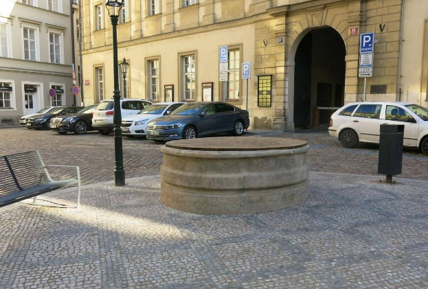 Ein Stadtplatz mit einer zentralen Bank, umgeben von Gebäuden mit Fenstern, Laternen und Schildern, mit einem Springbrunnen in der Mitte.