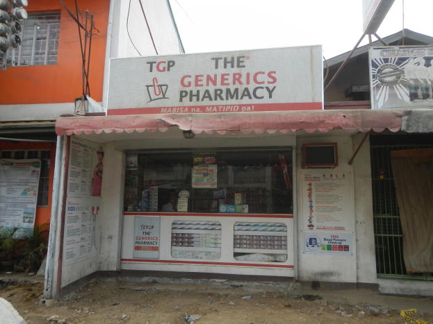 Eine Apotheke namens "Die Generika Apotheke" in einer urbanen Umgebung mit sichtbarer Infrastruktur und Fahrzeugen unter einem klaren Himmel.