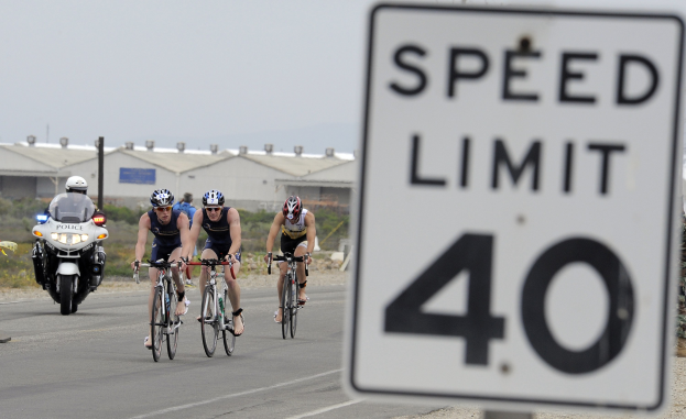 Eine Gruppe von Radfahrern mit Helmen fährt auf einer Straße mit einem Geschwindigkeitsbegrenzungsschild, Gebäude und Bäume im Hintergrund unter einem klaren blauen Himmel.
