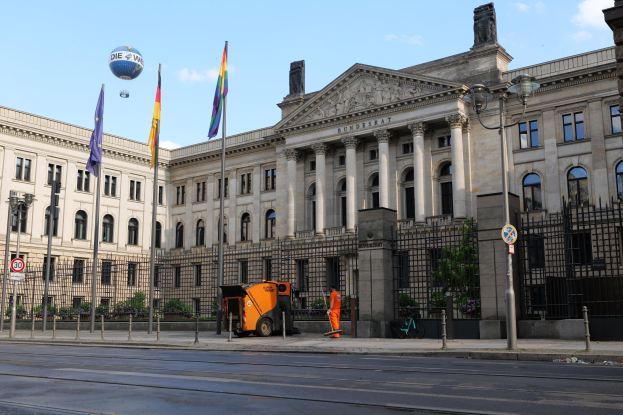 Großes modernes Gebäude mit vielen Fenstern, als Bundestag in Berlin identifiziert, umgeben von Straßeninfrastruktur, Fahrzeugen, Fußgängern, Flaggen und Bäumen unter einem bewölkten Himmel.