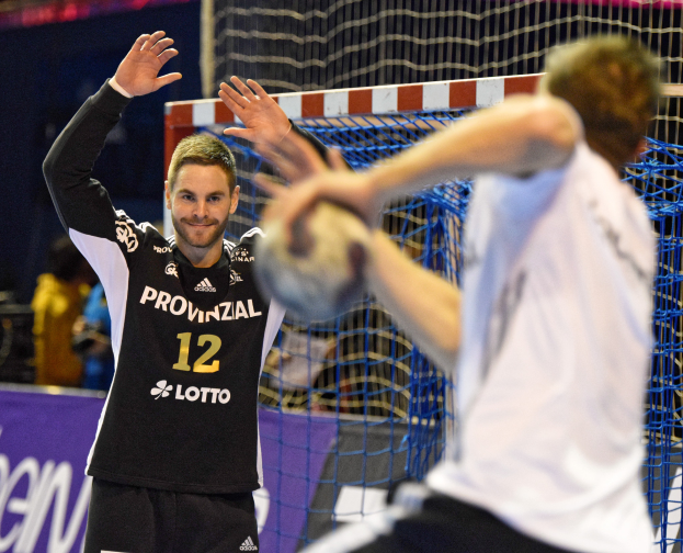 Zwei Männer beim Handballspielen vor einem Tor mit Netz und einem Banner mit Text im Hintergrund sowie einigen Zuschauern in einer Stadion.