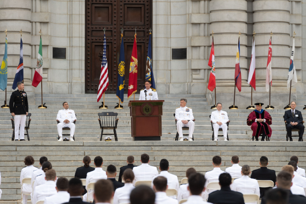 Gruppe von Menschen in weißen Uniformen auf den Stufen eines Gebäudes mit einem Podium, das von Fahnen flankiert ist, vor einem großen Gebäude mit einer Tür während einer feierlichen Zeremonie.