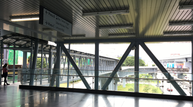 Innenansicht eines Flughafen-Terminals mit Menschen, Geländern, Säulen, Informationsanzeigen, Deckenleuchten und Außenelementen wie Straße, Brücke, Bäumen, Gras, Gebäuden und Himmel durch Fenster sichtbar.