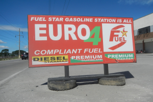 Eine Tankstelle mit einem "Euro4 Fuel"-Schild im Vordergrund, Fahrzeuge auf der Straße, Strommästele mit Drähten, Bäume und ein Gebäude im Hintergrund, der Himmel ist sichtbar.