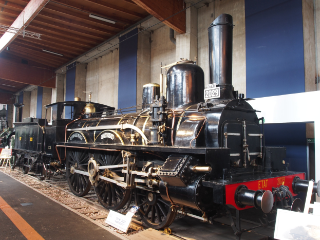 Eine große, alte Dampflokomotive in einem Museums Setting umgeben von Eisenbahnschienen, Informationsschildern und anderen Ausstellungen, mit einer beleuchteten Decke und Wand im Hintergrund.