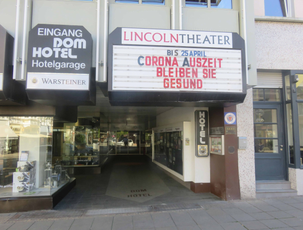 Außenansicht des Lincoln Theaters in Berlin mit Glasfenstern und -türen sowie einer Tafel mit Text und einem belebten Stadtbild im Inneren.