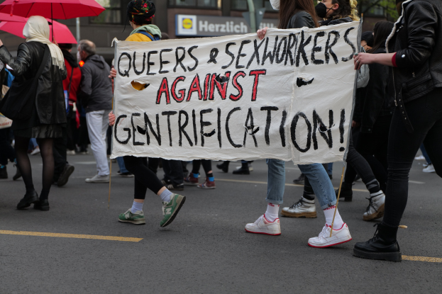 Gruppe von Menschen marschiert mit einem Banner 'Queers und Sexworker gegen Gentrifizierung' die Straße entlang, einige tragen Masken, einer hält einen Schirm, mit Bäumen und einem Gebäude mit Fenstern im Hintergrund.
