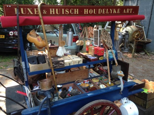 Ein blauer Karren voller Flaschen und Kisten auf einem Schotterweg auf einem Trödelmarkt, mit Fahrzeugen, Bäumen und einer Wand im Hintergrund.