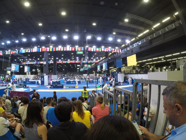 Eine große Menschenmenge sitzt und steht in einer Indoor-Sportarena, mit einem Metallzaun auf der rechten Seite, Bildschirmen, Flaggen und Deckenlampen im Hintergrund.