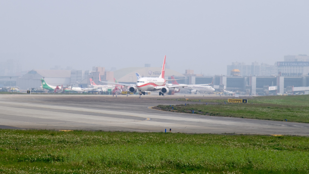 Flughafenrollbahn mit mehreren geparkten Flugzeugen, grasigem Umfeld, Gebäuden im Hintergrund und einem klaren blauen Himmel.