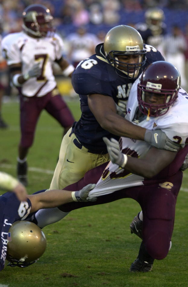 Zwei Fußballer in Helmen und Uniformen stoßen auf dem Feld zusammen, während andere zuschauen.