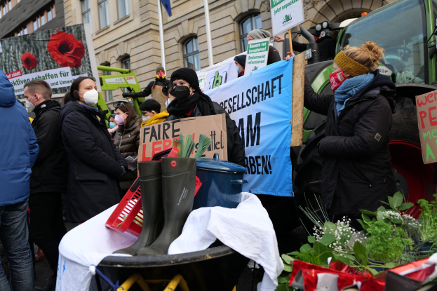 Eine Gruppe von Menschen mit Masken und Protestschildern vor einem Lastwagen, mit einem Tisch, Pflanzen und Gebäuden im Hintergrund.