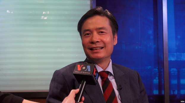 Ein Mann in Anzug und Krawatte hält ein Mikrofon, mit der Hand eines Reporters links daneben, spricht in einer Innenumgebung nahe einem Fenster.