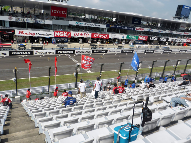 Eine Gruppe von Menschen sitzt auf der Tribüne einer Rennstrecke, umgeben von Fahnen, Schildern, Geländern, Stufen und anderen Gegenständen, sowie einem Stadion mit bewölktem Himmel im Hintergrund.