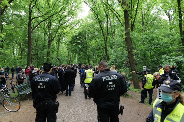 Polizisten vor einer Menschenmenge bei einer Anti-Terror-Demonstration in Berlin, mit Fahrrädern und einer Bank im Vordergrund und Bäumen im Hintergrund.
