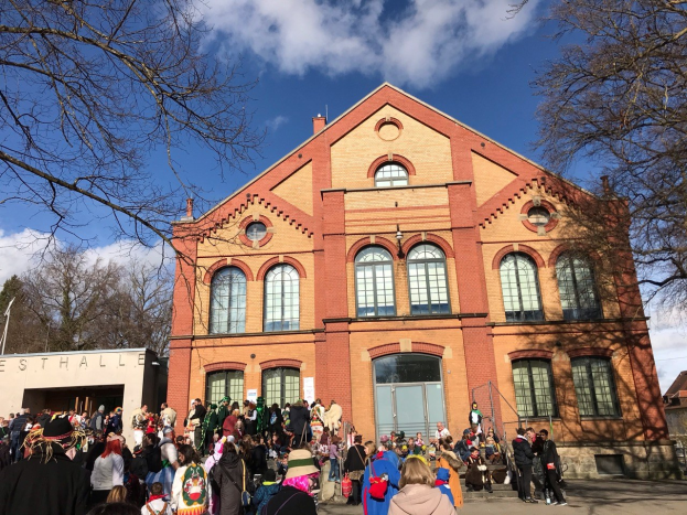 Eine große Gruppe von Menschen steht vor einem Gebäude mit Fenstern und Türen im Freien, umgeben von Bäumen unter einem bewölkten Himmel, einige tragen Mützen und Taschen.