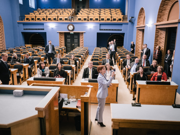 Frau hält Rede im schottischen Parlament, vor sitzenden und stehenden Zuhörern, Tischen mit Mikrofonen, Gläsern, Papieren und anderen Gegenständen, Uhr an der Wand und Deckenbeleuchtung im Hintergrund.
