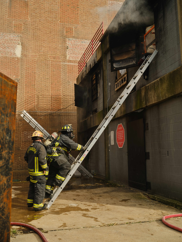 Feuerwehrleute mit Helmen klettern eine Leiter zu einem raucherfüllten Gebäude hinauf, mit einem Metallobjekt links und Rohren an der Basis.