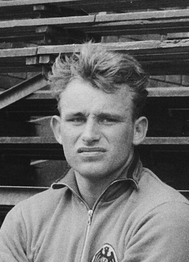 Schwarzes und weißes Foto eines Mannes in einer Jacke, der vor gestapelten Holzplanken steht, der als legendärer deutscher Füßballer gilt.