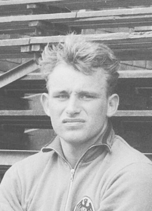 Schwarzes und weißes Foto eines Mannes in einer Jacke, der vor gestapelten Holzplanken steht, der als legendärer deutscher Füßballer gilt.