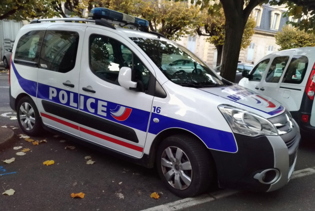Ein Polizeiwagen, der auf der Seite einer Straße mit Bäumen und Gebäuden im Hintergrund geparkt ist und ein sichtbares Vorderrad zeigt.