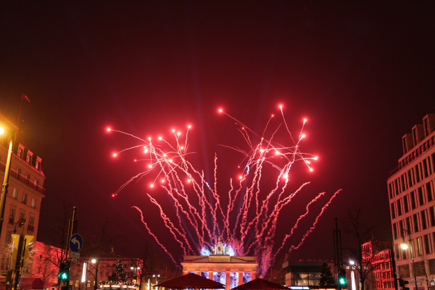 Eine Stadtstraßenszene mit Gebäuden, Bäumen, Laternen, Verkehrsschildern, Schildern, Zelten und Menschen vor einem Nachthimmel mit Feuerwerk während einer Silvesterfeier in Berlin.