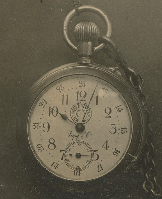 Old black and white photo of a pocket watch with a chain, featuring a numbered dial.