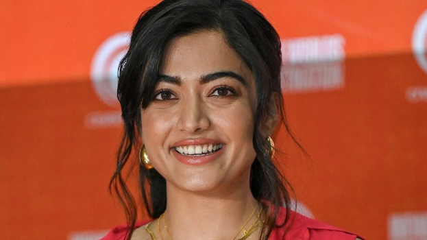 Eine Frau in einem roten Kleid mit einem eleganten Dutt und natürlichem Make-up lächelt in die Kamera, trägt schwarze High Heels und hat ein hellgelbes Banner mit der Aufschrift 'Sonam Kapoors beste Schönheits Looks vom Cannes Film Festival' im Hintergrund.