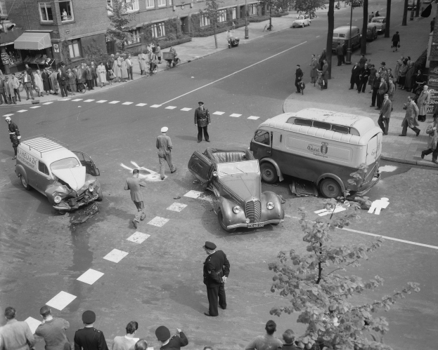 Schwarz-weiß-Szene eines Autounfalls an einer Straßenkreuzung mit mehreren Fahrzeugen, darunter ein Van, und einer Gruppe von Zuschauern, mit Gebäuden, Bäumen, Strommasten und Texttafeln im Hintergrund.