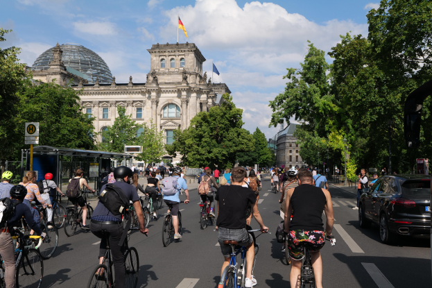 Eine Gruppe von Menschen fährt Fahrräder auf einer von Bäumen gesäumten Straße in Berlin, Deutschland, mit Gebäuden und einer Bushaltestelle, unter einem bewölkten Himmel und einer Flagge auf einem Gebäude.
