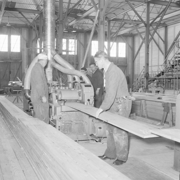 Schwarzes und weißes Foto von Männern, die an einer Maschine in einer mit Holzplanken, Pfählen, Lampen und Fenstern vollgestellten Fabrik arbeiten.
