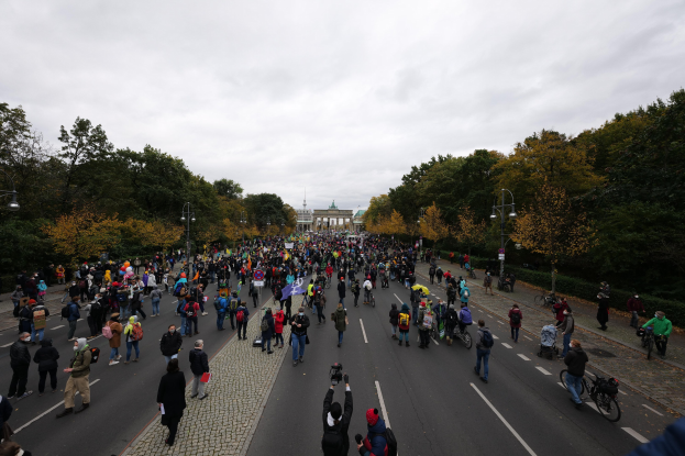 Große Gruppe von Menschen, die eine von Bäumen gesäumte Straße mit Laternenpfählen entlanggehen und Kameras halten, während sie an einer Protestdemo in Berlin teilnehmen, mit einem Gebäude und einem klaren Himmel im Hintergrund.