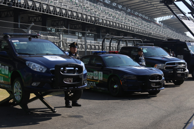 Polizeiautos vor einer Rennstrecke mit uniformierten Personen, ein Stadion mit Gelönd und Maschendrahtzaun sowie ein Himmel im Hintergrund.