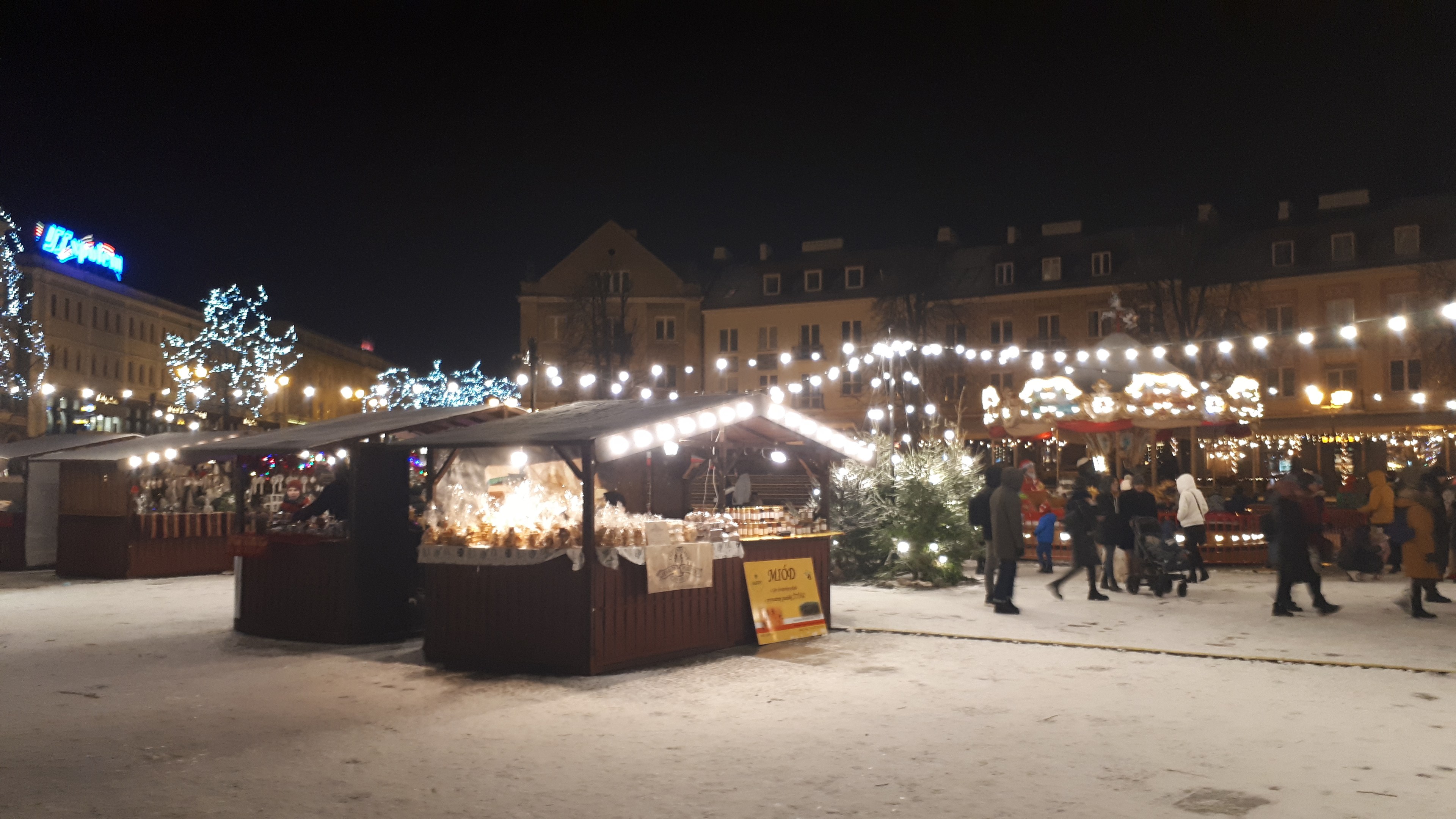 Ein nächtlicher Weihnachtsmarkt auf einer verschneiten Straße mit Menschen, beleuchteten Hütten, Pflanzen, Bäumen, Gebäuden und Schildern unter einem bewölkten Himmel.
