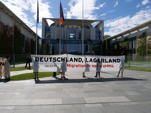 Gruppe von Menschen mit einem Banner, auf dem "Deutschland, Lagerland Migration ist kein Verbrechen" steht, vor einem umzäunten Gebäude mit Bäumen und Fahnen.