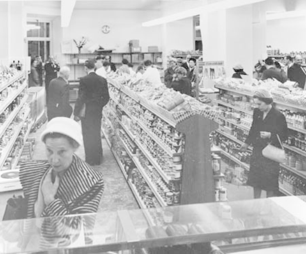 Schwarzes und weißes Foto von Menschen beim Einkaufen in einem Supermarkt mit Lebensmitteln auf Regalen, einige halten Taschen, eine Uhr an der Wand und Deckenbeleuchtung.