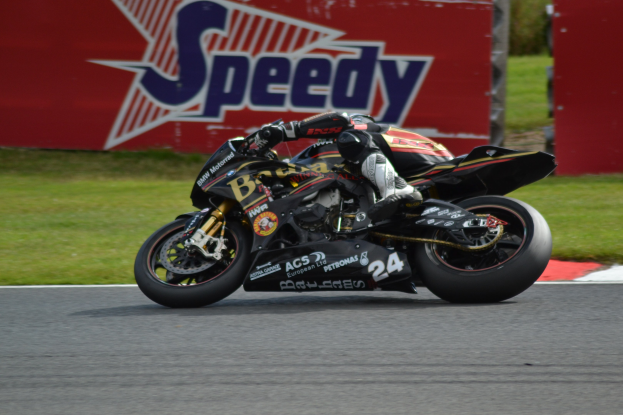 Eine Person in Helm und Schuhwerk fährt ein Motorrad auf einer Rennstrecke, umgeben von Gras und einer Tafel mit Text im Hintergrund.