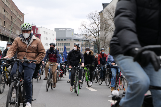 Gruppe von Radfahrern in Helmen und Handschuhen auf einer von Bäumen gesäumten Straße in Berlin fahrend, mit Gebäuden und einem geparkten Fahrzeug im Hintergrund.