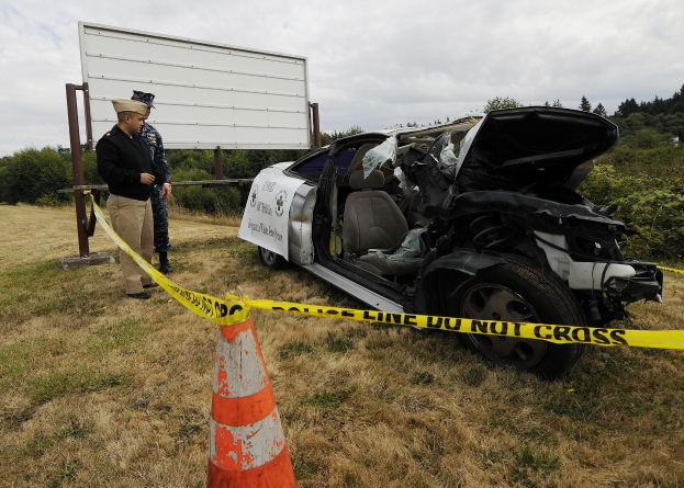 Ein Polizeibeamter in Uniform steht neben einem schwer beschädigten Auto am Straßenrand, mit eingedellter Motorhaube und zersplitterten Fenstern.