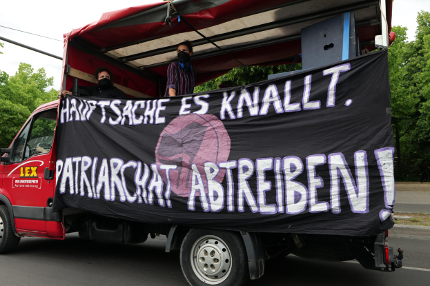 Roter Lastwagen mit einer Fahne fährt eine von Bäumen gesäumte Straße unter einem klaren blauen Himmel entlang, zwei maskierte Personen sind im Inneren sichtbar.