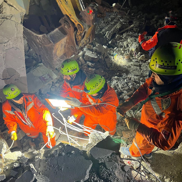 Gruppe von Menschen in orangen Uniformen und grünen Helmen bei der Arbeit an Trömmern eines eingestürzten Gebäudes mit einem Kran im Hintergrund.