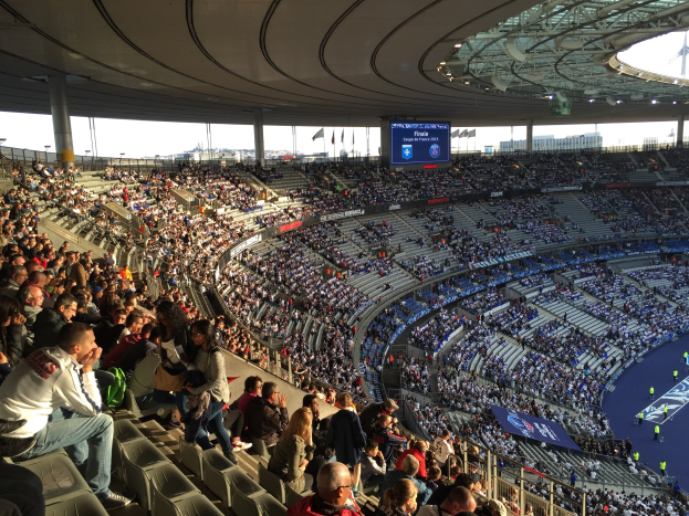 Große Menschenmenge in einem Stadion bei einem Fötballspiel, mit einer Bühne rechts, Fahnen, Stangen, einem Bildschirm und der Allianz Arena in München, Deutschland im Hintergrund.